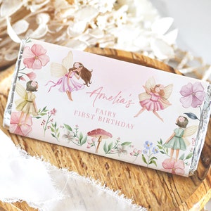 May include: A chocolate bar wrapper with a pink and white gradient. It features illustrations of fairies, flowers, and the text "Amelia's Fairy First Birthday". The design incorporates pink, green, and brown colours.
