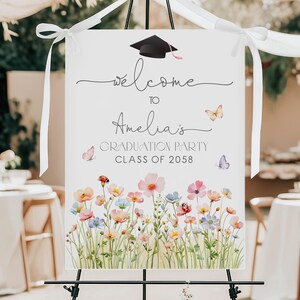 May include: A white graduation party welcome sign with the text "Welcome to Amelia's Graduation Party Class of 2058." The sign features a black graduation cap, butterflies, and a watercolor floral design with pink, blue, and yellow flowers.