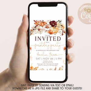 May include: A smartphone displays a digital invitation for a first birthday party. The invitation features a floral arrangement with pumpkins and fall foliage. Text includes the event details and the phrase "You're Invited".