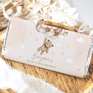 May include: A pink and brown chocolate bar wrapper featuring a teddy bear holding onto balloons. The wrapper has the text "Benjamin's Bear-y First Birthday" printed on it.