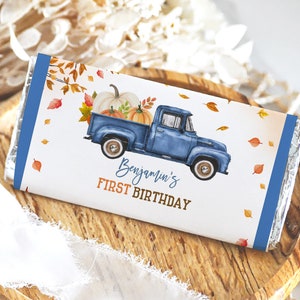 May include: A blue and white truck with a bed full of pumpkins and fall leaves. The truck has a blue cab and a white bed. The truck is on a white background with blue and orange accents. The text "Benjamin's First Birthday" is printed on the wrapper.