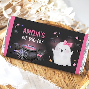 May include: A pink and black Halloween-themed chocolate bar wrapper with a ghost, a cauldron, a black cat, and the text "Amelia's 1st Boo-Day".