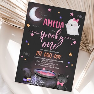 May include: A black and white invitation card with a pink and purple design. The card says "Amelia is a spooky one" and "1st Boo-Day". The card also features a ghost, a cauldron, and a black cat.