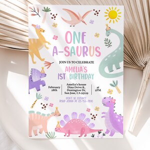 May include: A colorful invitation for a first birthday party with a dinosaur theme. The invitation features a variety of dinosaurs in pink, purple, blue, and yellow, with the text "ONE A-SAURUS" in pink and purple letters. The invitation also includes the date, time, and location of the party.