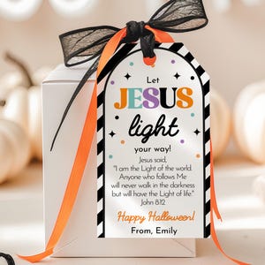 Op de afbeelding: Een witte geschenkdoos met een zwart en oranje lint en een decoratief label. Het label bevat de tekst "Let JESUS light your way!" en "Happy Halloween!". Witte pompoenen zijn zichtbaar op de achtergrond.