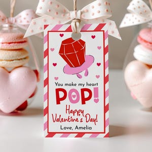 May include: Valentine's Day gift tag with a red and pink design. The tag features a cartoon ring and the text "You make my heart POP! Happy Valentine's Day! Love, Amelia." The tag is tied with a white ribbon to a clear container filled with macarons and a heart-shaped candy.