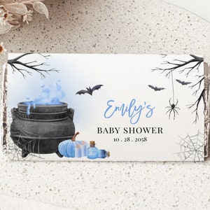 May include: A white chocolate bar wrapper with a Halloween theme. The wrapper features a black cauldron with blue smoke, a spider web, bats, and a pumpkin. The text on the wrapper reads "Emily's Baby Shower 10.28.2058".