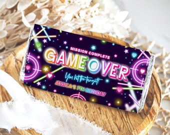 Puede incluir: Un envoltorio de chocolate Game Over colorido con luces de neón, estrellas y corazones sobre un fondo morado oscuro. El texto dice "Misión Completa Game Over ¡Has dado en el blanco! Jessica's 7th Birthday".