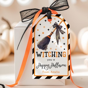 May include: A white gift box with a Halloween-themed tag. The tag features a watercolor broom illustration and the text "Witching you a Happy Halloween". It is adorned with a black ribbon and orange ribbon.