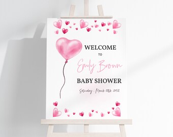 May include: A white welcome sign with pink hearts and a pink heart-shaped balloon. The sign reads "Welcome to Emily Brown Baby Shower Saturday, March 25th, 2058".