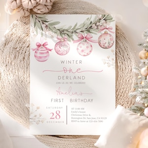 May include: A rectangular invitation card featuring a watercolour illustration of a green pine garland with four hanging pink Christmas ornaments. The text reads: WINTER ONE DERLAND, JOIN US AS WE CELEBRATE Amelia's FIRST BIRTHDAY, SATURDAY 28 DECEMBER, AT 11:00 AM, Emily's house, Christmas Drive &, Dunnington Dr, San Jose, CA 95129, RSVP to Anne at 123-456-7890.