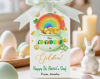 May include: A white gift tag with a green ribbon features a pot of gold under a rainbow, with the text "Our Friendship is Golden!" and "Happy St. Patrick's Day!" A white ceramic bunny and candles are in the background.
