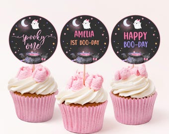 Op de afbeelding: Drie roze en witte cupcakes met zwarte en witte Halloween-toppers. De toppers tonen een spook, een ketel en de tekst "spooky one", "Amelia is Boo-Day" en "Happy Boo-Day".