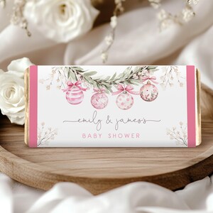 May include: A rectangular candy bar wrapper with pink accents and gold foil edges. The wrapper features pink and white ornaments, greenery, and the text "emily & james's BABY SHOWER" in a delicate script.
