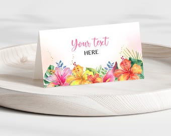 Peut inclure: Une carte blanche pliée avec le texte "Your text HERE" en rose et noir. La carte est décorée de fleurs à l'aquarelle dans les tons rose, jaune et orange, avec des feuilles vertes. La carte est posée sur un plateau en bois blanc.