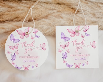 May include: Two white tags with butterfly designs. One is round, the other square. Both have pink and purple watercolor butterflies and the text "Thank you for Fluttering By! Love, Amelia" in pink script.