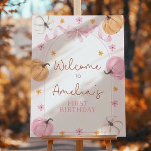 Puede incluir: Un letrero de bienvenida para una celebración de primer cumpleaños. El letrero presenta calabazas de acuarela en tonos rosa, blanco y naranja, junto con detalles florales y una cinta rosa. El texto dice "Welcome to Amelia's First Birthday".
