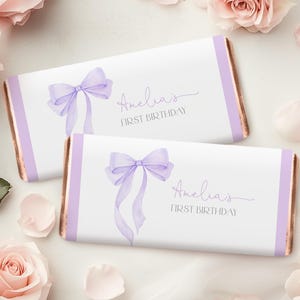 May include: Two white candy bar wrappers featuring a lavender watercolour bow and the text "Amelia's FIRST BIRTHDAY". The wrappers have lavender borders and a gold-coloured edge. Pink roses and petals surround the candy bars.