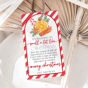 May include: A rectangular gift tag with a red and white striped border and a brown string. The tag features a watercolor illustration of an orange slice, cinnamon sticks, and greenery. Text on the tag reads "It's beginning to smell a lot like Christmas."