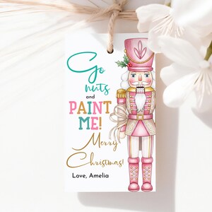 May include: A white gift tag with a pink and gold nutcracker illustration. The tag has the text "Go nuts and PAINT ME! Merry Christmas! Love, Amelia" in various colors and fonts. The tag is tied with twine.