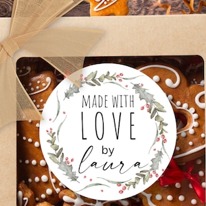 May include: A square cardboard gift box with a clear window, topped with a beige organza ribbon. Inside are gingerbread cookies. A circular white sticker features a green holly wreath and the text MADE WITH LOVE BY LAURA.