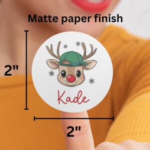 May include: A 2-inch round sticker with a matte finish featuring a cartoon reindeer wearing a green hat and a red nose. The sticker says "Kade" in red lettering.