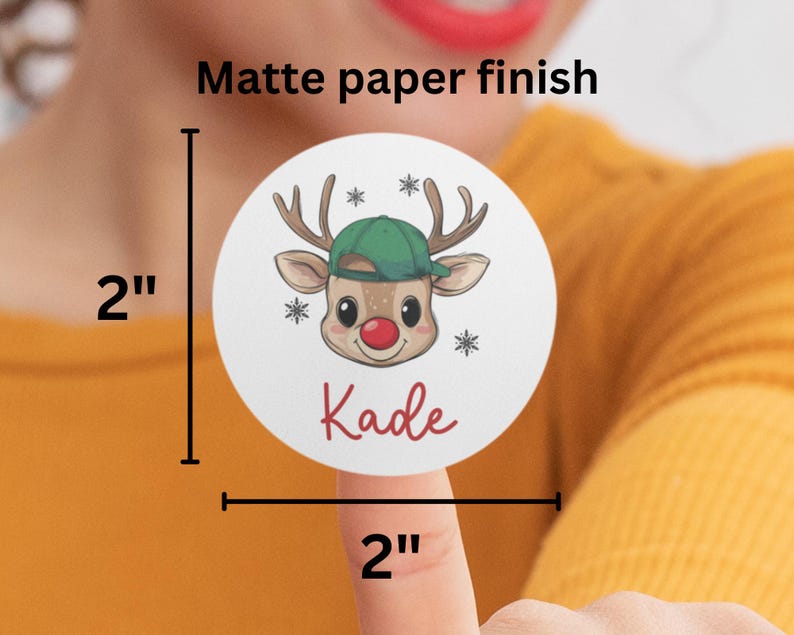 May include: A 2-inch round sticker with a matte finish featuring a cartoon reindeer wearing a green hat and a red nose. The sticker says "Kade" in red lettering.