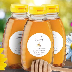 May include: Three bottles of pure honey with yellow caps and white labels. The labels read "pure honey" with a bee graphic and text indicating hand-harvesting. A wooden honey dipper rests in front of the bottles. The background includes flowers and greenery.