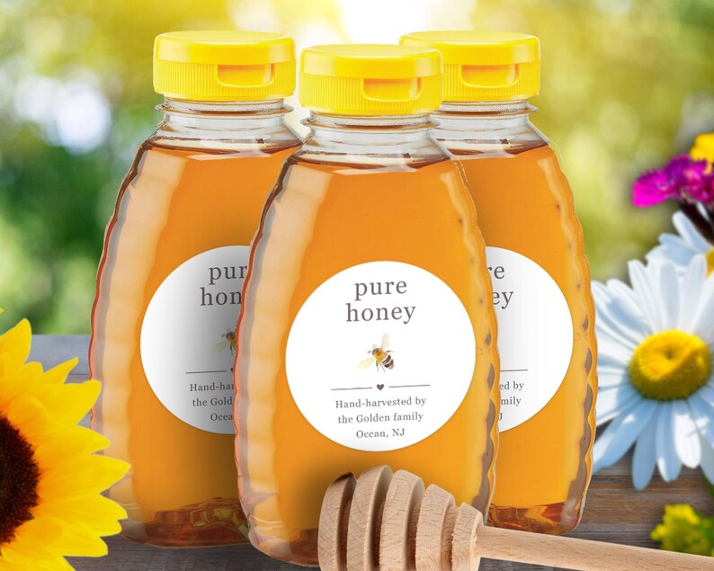 May include: Three bottles of pure honey with yellow caps and white labels. The labels read "pure honey" with a bee graphic and text indicating hand-harvesting. A wooden honey dipper rests in front of the bottles. The background includes flowers and greenery.