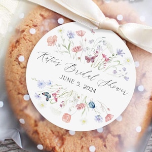 May include: A round white sticker with a watercolor floral design in shades of pink, blue, and green. The sticker says "Katie's Bridal Shower" with the date "June 5, 2024" underneath.