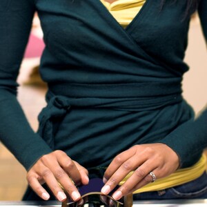 K&ouml;nnte beinhalten: Ein dunkel t&uuml;rkisfarbener Wickelpullover mit langen &Auml;rmeln, der in der Taille gebunden ist. Der Pullover ist &uuml;ber einem gelben Oberteil und einer blauen Jeans getragen. Die Person tr&auml;gt einen silbernen Ring. Die H&auml;nde halten ein dunkles Objekt.