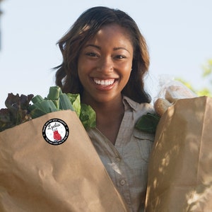 May include: A woman smiling while holding two brown paper grocery bags filled with groceries. One bag contains leafy greens, and the other contains bread. A round sticker on the bag reads "Thank you for your support! Lydia. You can rate me in the app!"