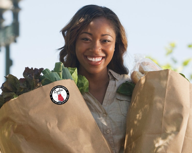 May include: A woman smiling while holding two brown paper grocery bags filled with groceries. One bag contains leafy greens, and the other contains bread. A round sticker on the bag reads "Thank you for your support! Lydia. You can rate me in the app!"