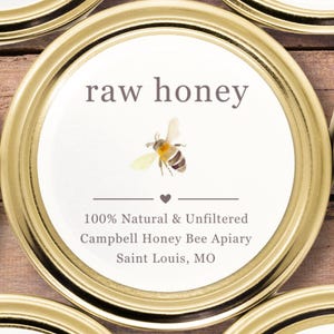 May include: Close-up of a jar lid with the words "raw honey" in a serif font. A watercolor illustration of a bee is centered below the text. The label also includes the text "100% Natural & Unfiltered" and the apiary's name and location.