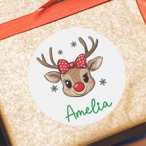 May include: A round sticker with a cartoon reindeer with red and white polka dot bow. The reindeer has a red nose and the sticker says "Amelia" in green text.