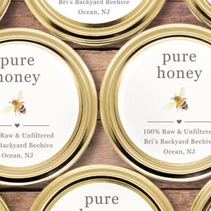 May include: Close-up of several golden jar lids with white labels. The labels read "pure honey" with a bee illustration. Text below states "100% Raw & Unfiltered" and the name and location of the beekeeper.