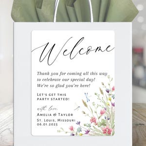 May include: A white welcome bag with a floral design and the text "Welcome, Thank you for coming all this way to celebrate our special day! We're so glad you're here! Let's get this party started! with love, Amelia & Taylor, St. Louis, Missouri, 06.01.2025"