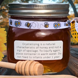 May include: A jar of honey with a silver lid and a white label. The label reads, "Crystallizing is a natural characteristic of honey..." A decorative ribbon with bees and flowers encircles the jar. A wooden honey dipper rests in front.
