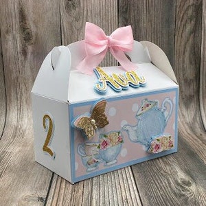 May include: A white favor box with a pink bow and a blue and white teacup design. The box has the name "Ava" and the number "2" on it.