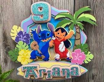 May include: A colorful cake topper featuring the characters Stitch and Lilo from the Disney movie Lilo & Stitch. The number 9 is displayed above the characters, and the name "Ariana" is written below. The design includes tropical flowers and a palm tree.