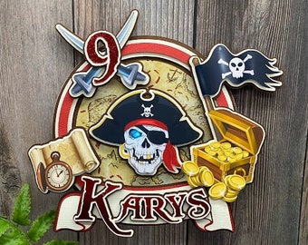Peut inclure: Un topper de gâteau d'anniversaire sur le thème des pirates avec un drapeau de crâne et d'os croisés, un chapeau de pirate, un coffre au trésor et le nom "KARYS" écrit dans une police décorative.