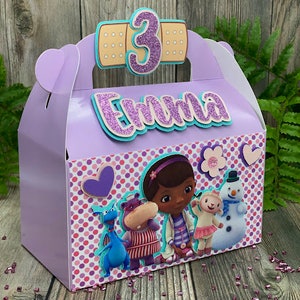 May include: A lavender party favor box with a handle, featuring a colorful design with cartoon characters and the name "Emma" in glittery letters. A large number "3" is on top, indicating a third birthday.