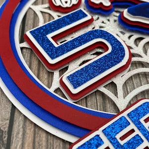 May include: A blue and red layered paper cake topper with a spider web design, a spider, and the number 5 and the word "Seven" in blue glitter.