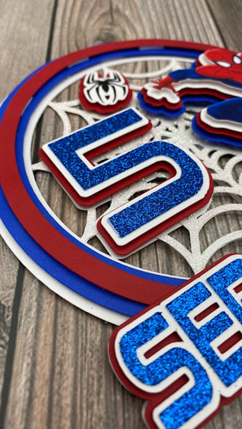 May include: A blue and red layered paper cake topper with a spider web design, a spider, and the number 5 and the word "Seven" in blue glitter.
