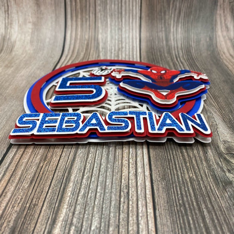 May include: A red, white and blue cake topper featuring a Spiderman character, the number 5, and the name "SEBASTIAN".