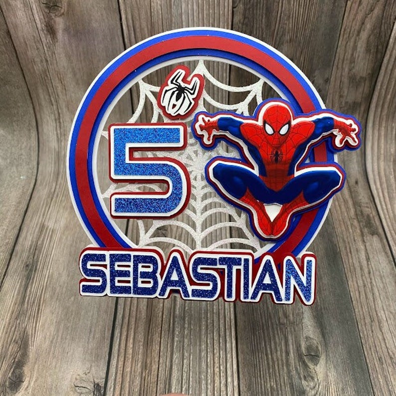 May include: A red, white and blue cake topper featuring a Spiderman figure, a spider web, the number 5, and the name Sebastian.