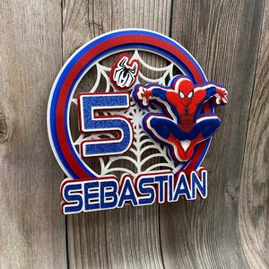 May include: A red, white and blue layered cake topper featuring a Spiderman character, a spider web, the number 5, and the name "SEBASTIAN".