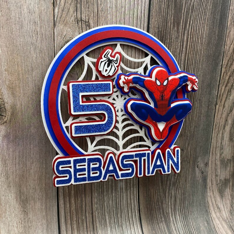 May include: A red, white and blue layered cake topper featuring a Spiderman character, a spider web, the number 5, and the name "SEBASTIAN".