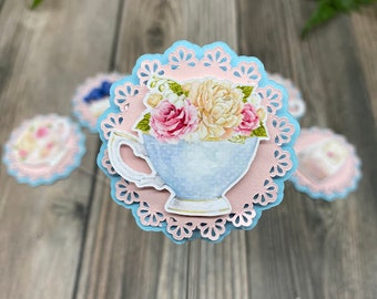 May include: A blue and pink cupcake topper with a delicate lace design. A blue teacup with a floral design is centered on the topper.