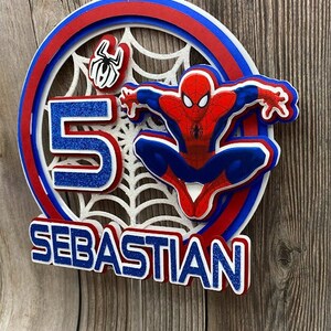 May include: A red, blue and white cake topper featuring Spiderman, a spiderweb and the number 5. The name Sebastian is written in blue glitter letters.